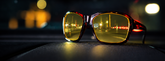 Close-up of tortoiseshell sunglasses with yellow-tinted lenses resting on a dark textured surface, with a blurred nighttime street scene reflected in the lenses.